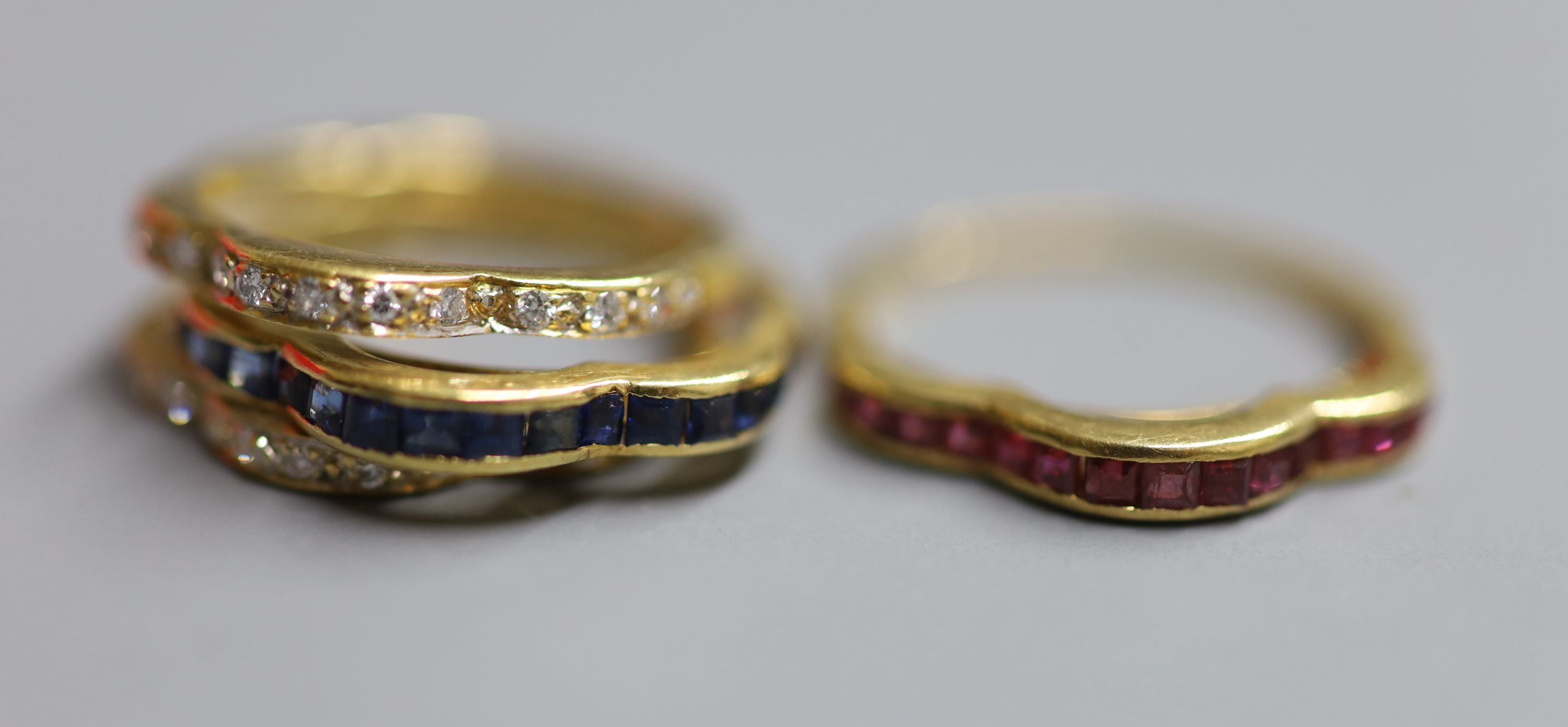 A modern continental 750 yellow metal and triple shank, sapphire, ruby and diamond set shaped half eternity ring,